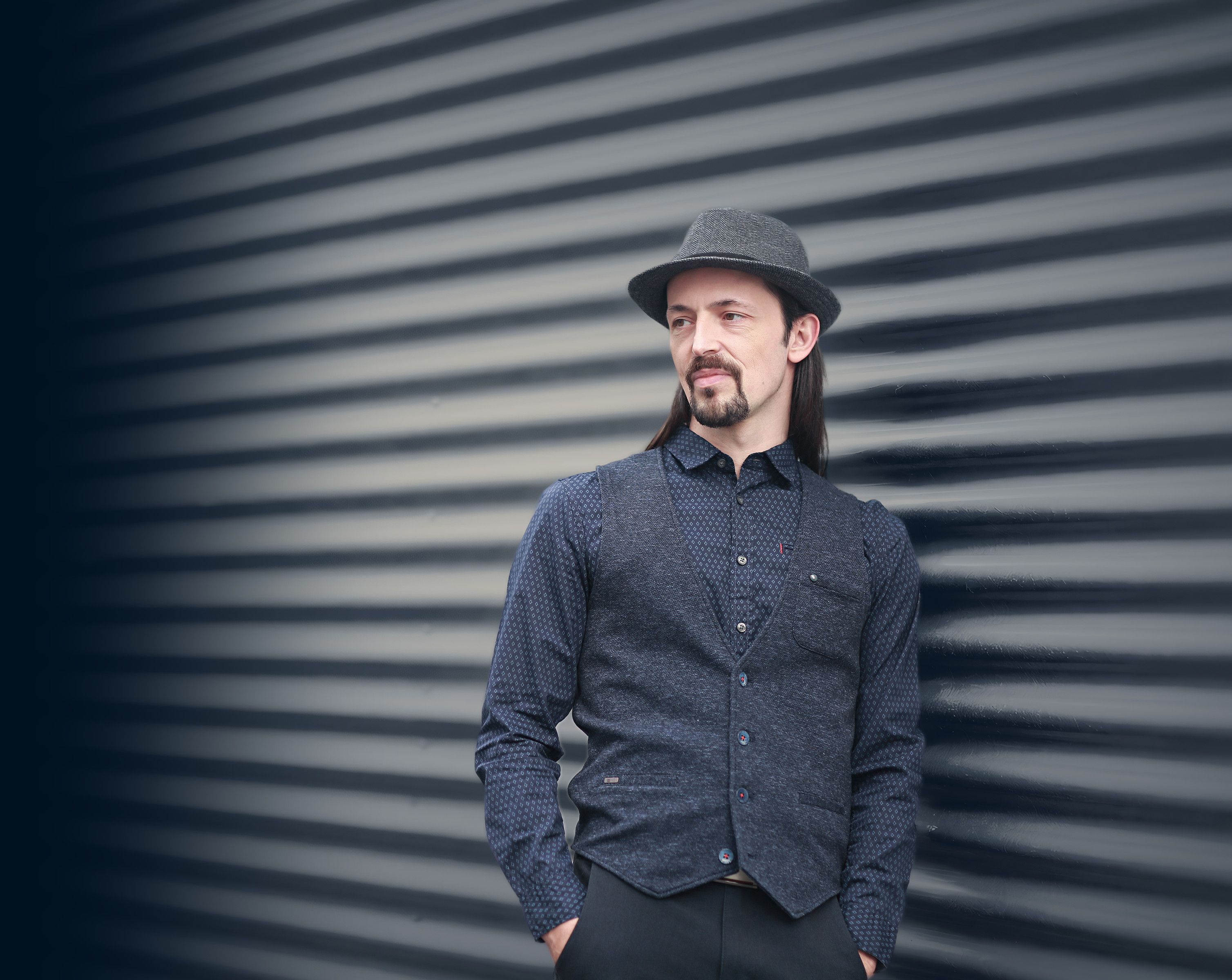 Alain Schlesser in a grey hat and navy waistcoat, leaning against a corrugated metal wall