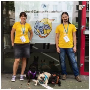 My wife Carole and me, with our dogs Jasper, Duke and Indra, in front of a WordCamp Frankfurt poster.